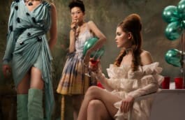 Three women in elegant dresses at a party. One in a blue dress with high boots, another in a pastel dress holding a green balloon, and the third in a white ruffled dress holding a glass of red liquid. 