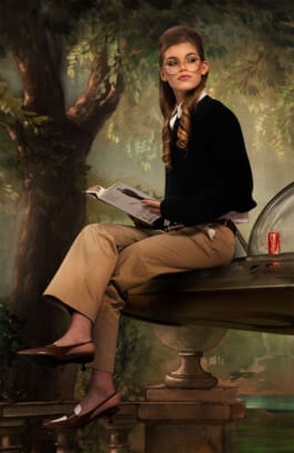 A woman with styled hair and glasses, dressed in a black sweater and beige pants, sitting on a metallic surface, reading a book with a backdrop of painted trees and a Coca-Cola can beside her.