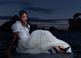 A woman in a white dress with puffy sleeves and a long skirt, sitting in water at dusk with rocky terrain in the background.